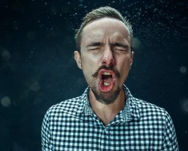 Young funny handsome man with beard and mustache sneezing with spray and small drops, studio portrait on black background(Master1305)s