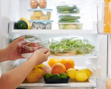 Hands of woman opening fridge door and putting package of fresh ripe strawberries in it