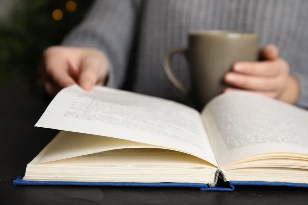 Woman,With,Cup,Of,Beverage,Reading,Book,At,Table,,Closeup