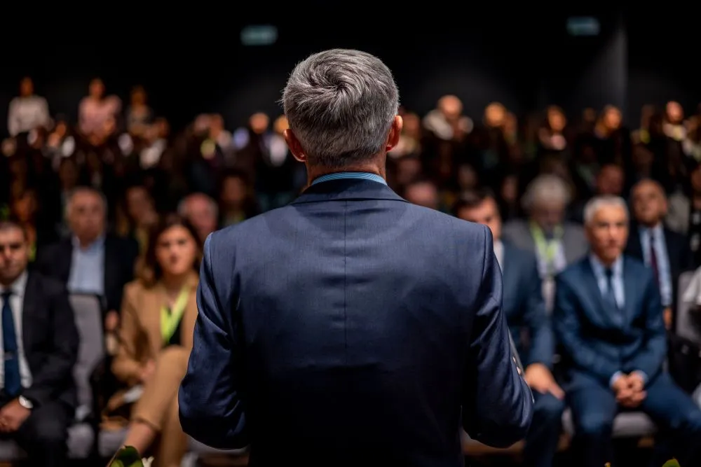 Business man is making a speech in front of a big audience at a conference hall. Speaker giving a talk on corporate business or political conference.